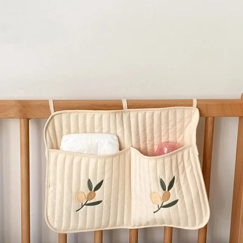 クリーム色のキルティング製ベビーカー用オーガナイザー袋（布製）で、刺繍のオリーブの枝モチーフと、赤ちゃんの必需品を入れるためのポケットが2つ付いています。