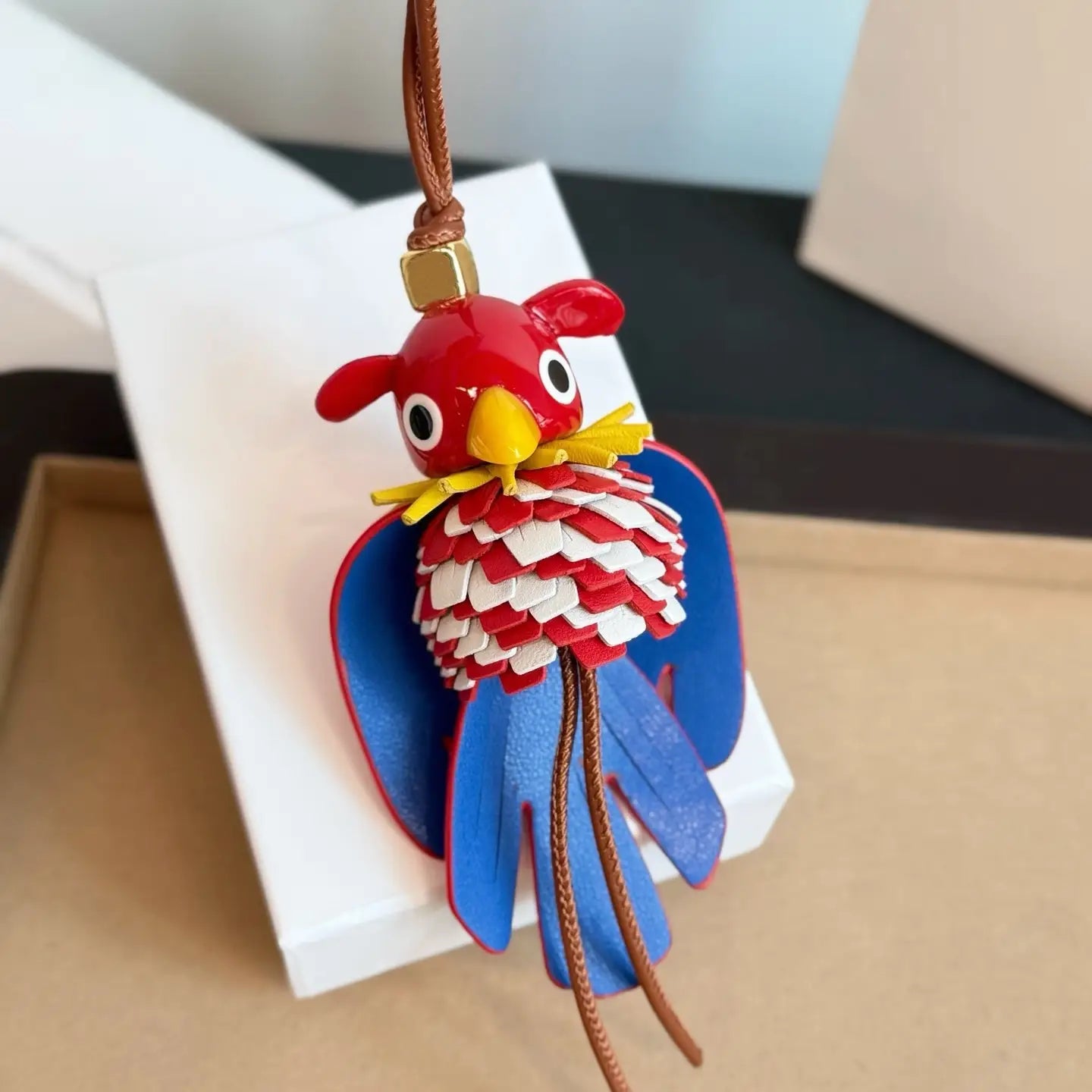 Un llavero colgante para bolso con encanto de animales de cuero con forma de loro rojo y azul vibrante, con pico amarillo y ojos blancos detallados, con diseño de plumas en capas.