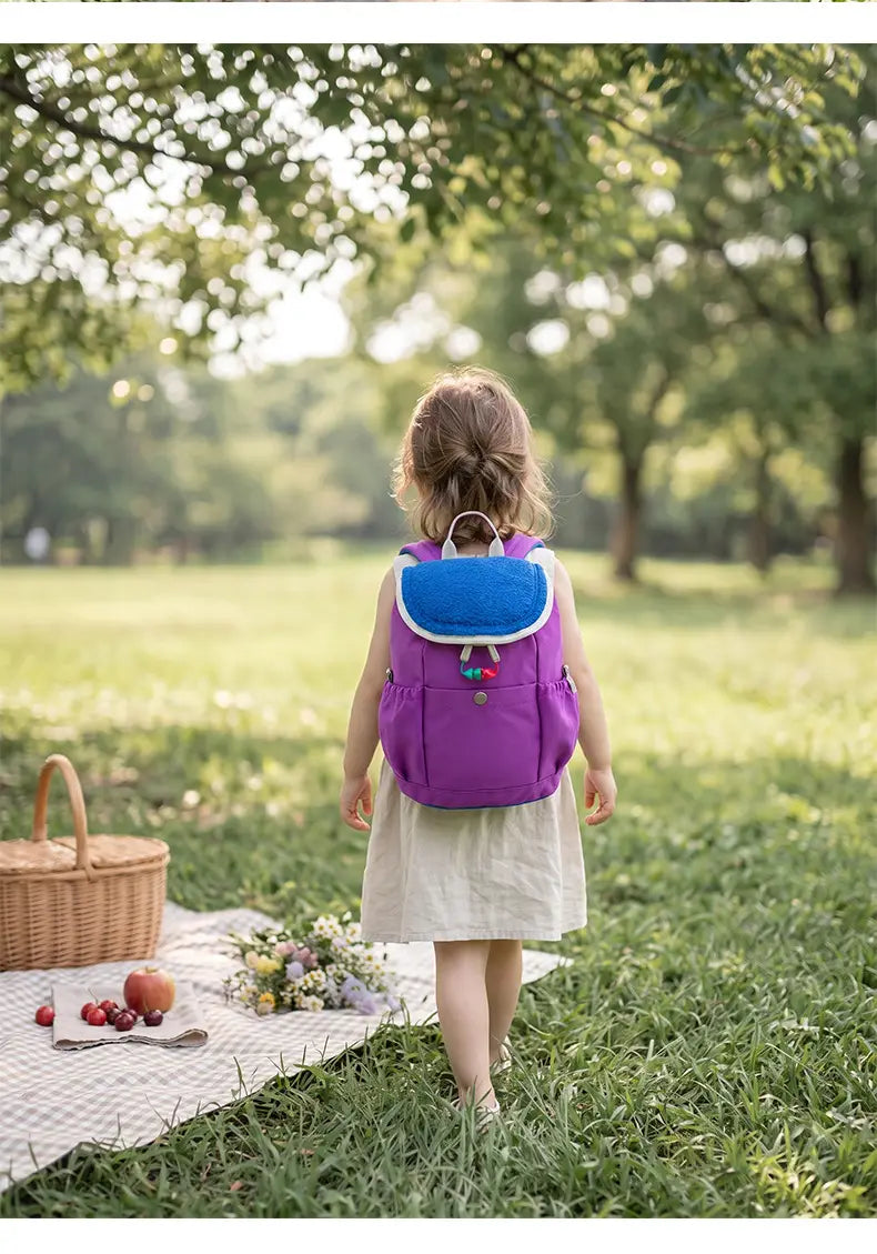 Leichter Fuzzy-Rucksack für Kinder in Lila mit einer leuchtend blauen vorderen Klappe und farbenfrohen Quasten, entworfen als Kindergartentagesrucksack.