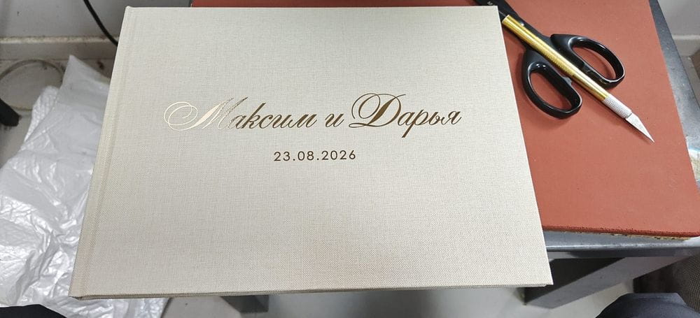 Cream-colored leather-bound custom wedding guest book and pen linen photo album with gold-embossed Cyrillic script and date on the cover.