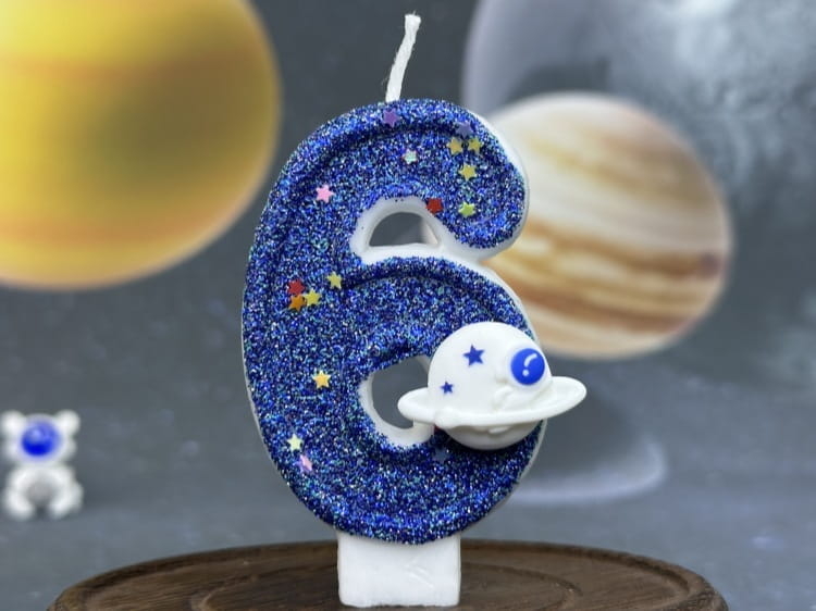 Una vela azul con brillo con el número seis para cumpleaños, con estrellas de colores y una pequeña figura de planeta blanco, diseñada como adorno para pastel de fiesta planetaria.