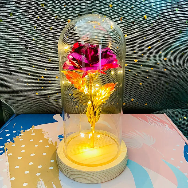 Eine leuchtende metallische Rose mit tiefem Magenta-Blütenkelch und goldenem Stiel, umschlossen in einer klaren Glaskuppel auf einem Holzsockel, LED-Licht-Geschenk.