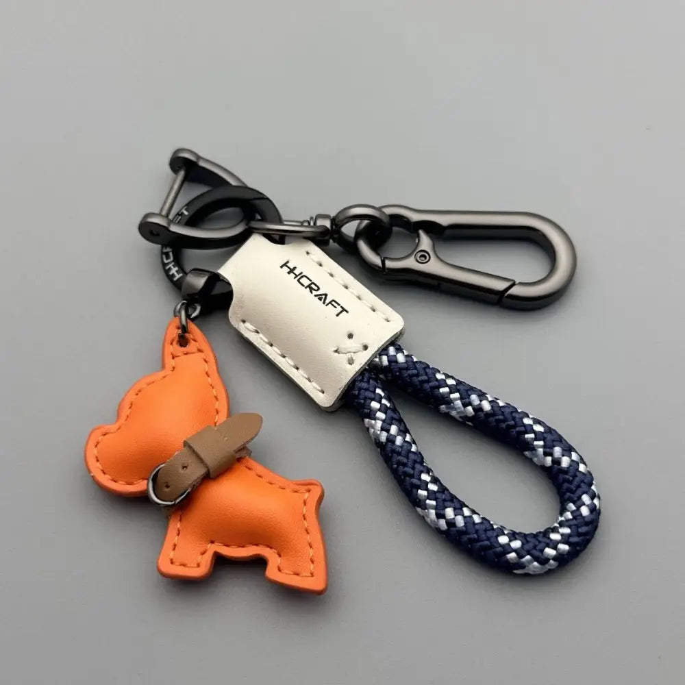 Orange leather French bulldog keychain with tan belt detail attached to a navy and white braided rope loop and metal hardware