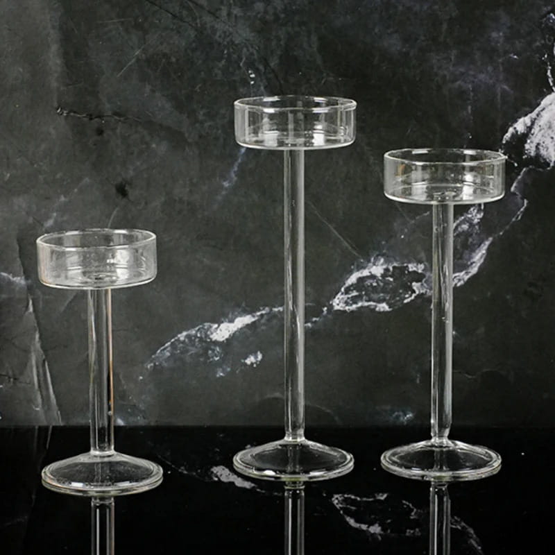 Tres portavelas de pilar de vidrio transparente, con partes superiores cilíndricas y tallos esbeltos de alturas variables, para una decoración moderna de mesa sobre un fondo de mármol oscuro.
