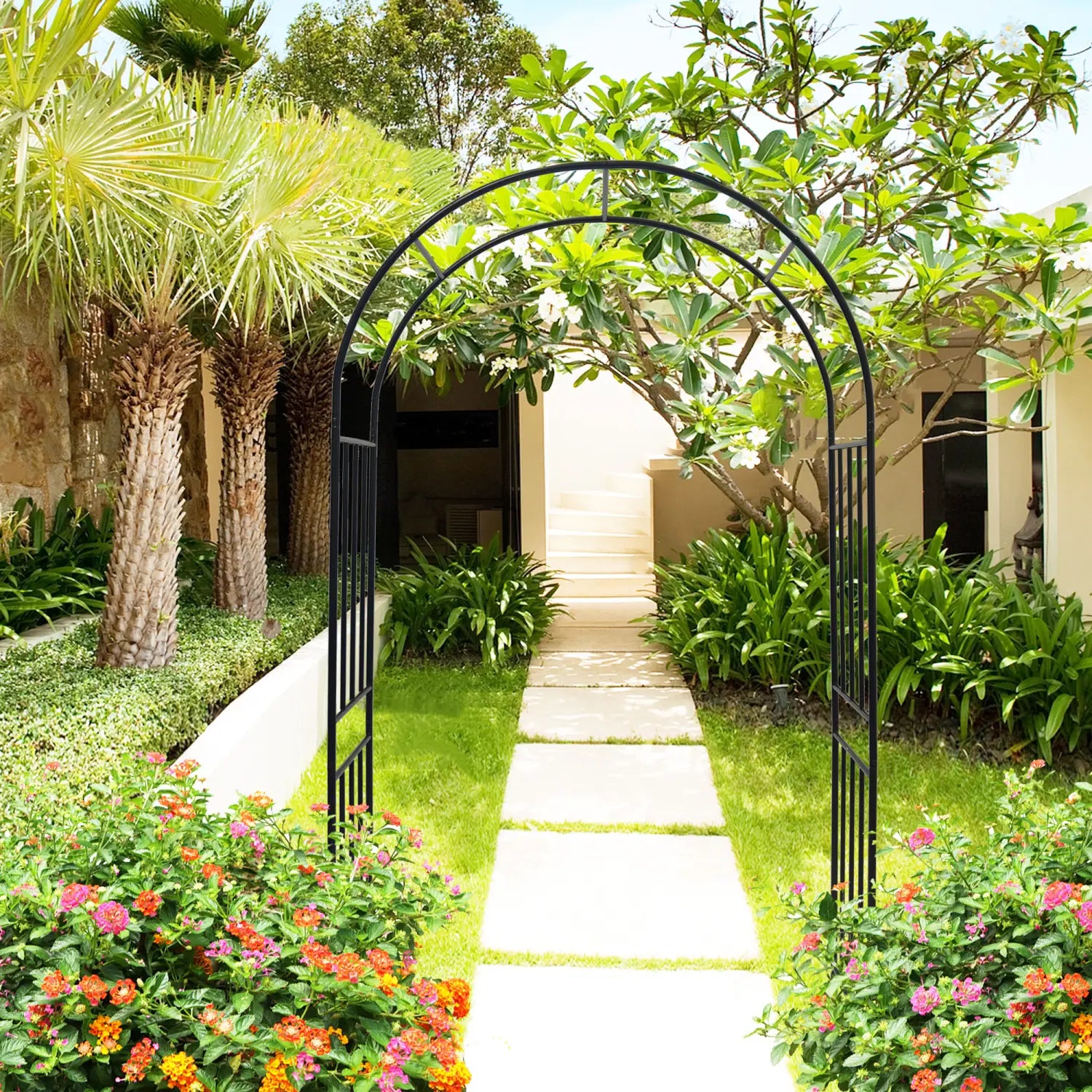 Black wrought iron metal garden arch with vertical slats framing a pathway lined by vibrant flowers and tropical plants.