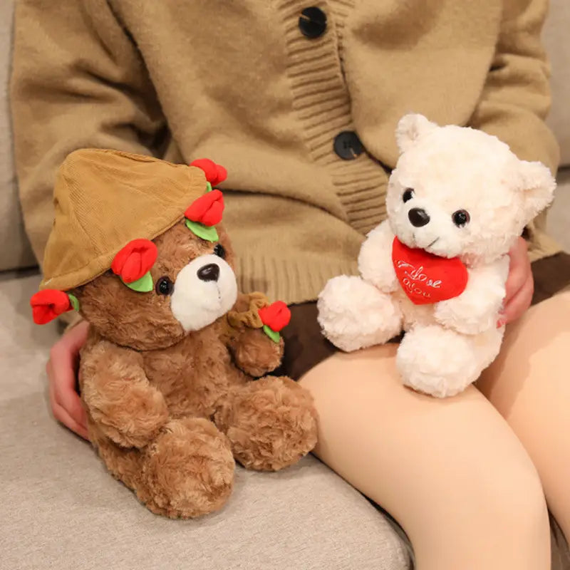 Ein brauner Plüsch-Teddybär trägt eine beigen Kordmütze, die mit roten Rosen verziert ist, und hält eine kleine Rose; neben einem weißen Stoffbären, der ein rotes Herz mit sich trägt, in einem Teddybär-Plüschtier mit Rosen, Valentinsgeschenk, in der Größe 16 bis 25 Zentimeter.