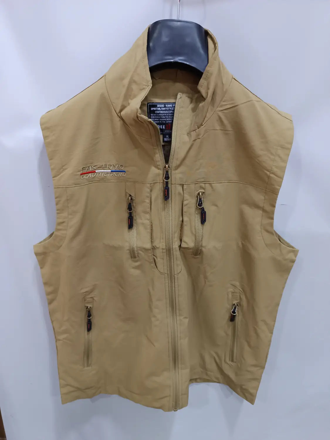 Chaleco utilitario con capucha y múltiples bolsillos para hombre en material impermeable color arena, con varios bolsillos con cremallera y un logotipo de la marca bordado en el lado izquierdo del pecho.