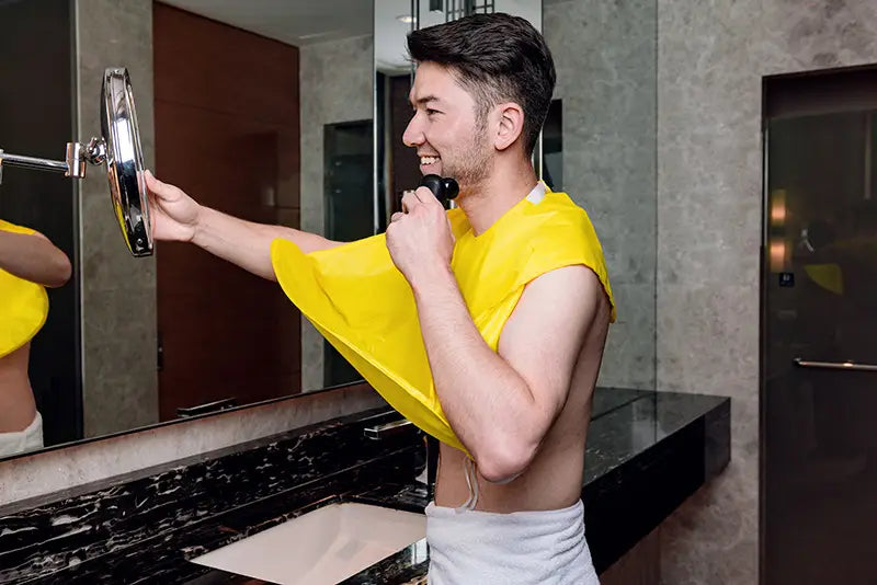 Ein Mann trägt einen leuchtend gelben, faltbaren, wasserdichten Rasier-Latz, einen Friseur-Cape-Schürzen, über die Schultern drapiert, mit weißen Handtüchern um die Taille, hält einen elektrischen Rasierer und richtet einen Vergrößerungsspiegel mit silbernem Rahmen aus.