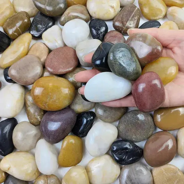 A hand holds a collection of polished river rocks in various earthy tones including white, black, brown, yellow, and gray for decorative use.