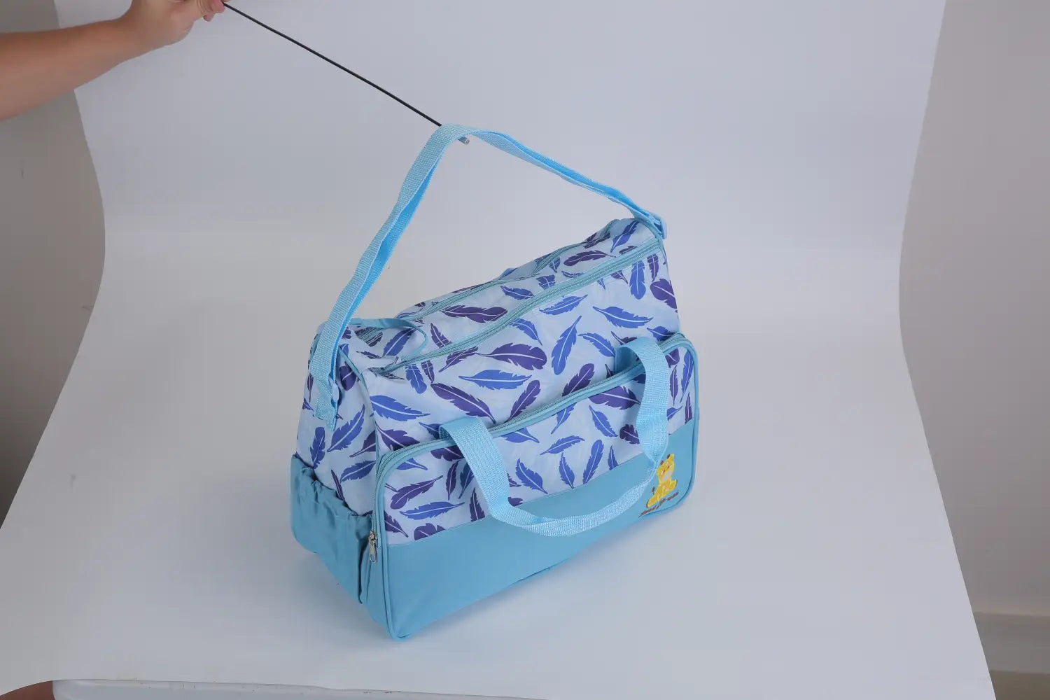Light blue baby diaper bag with a white base and purple feather patterns featuring a small yellow duck appliqué included in a 5 piece patterned baby diaper bag set with travel accessories.