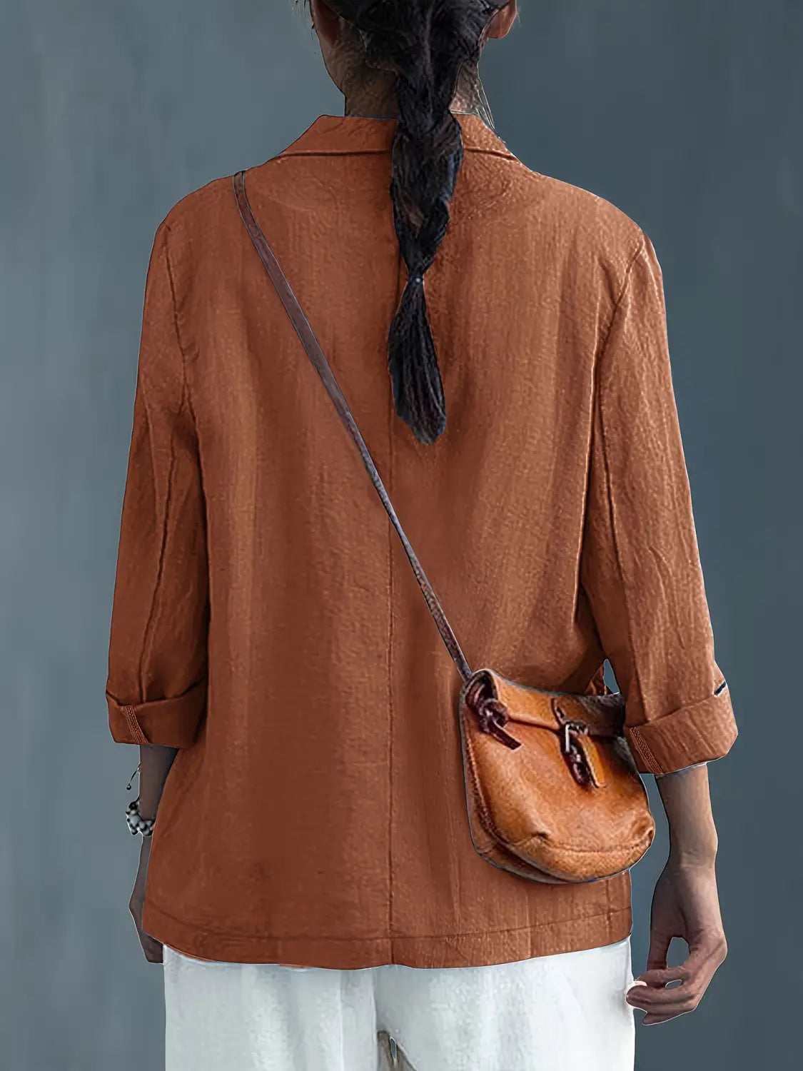 Un bolso cruzado pequeño de cuero color camel, con forma redondeada y dos cierres con hebilla, combinado con un blazer de lino para mujer, casual y de trabajo.