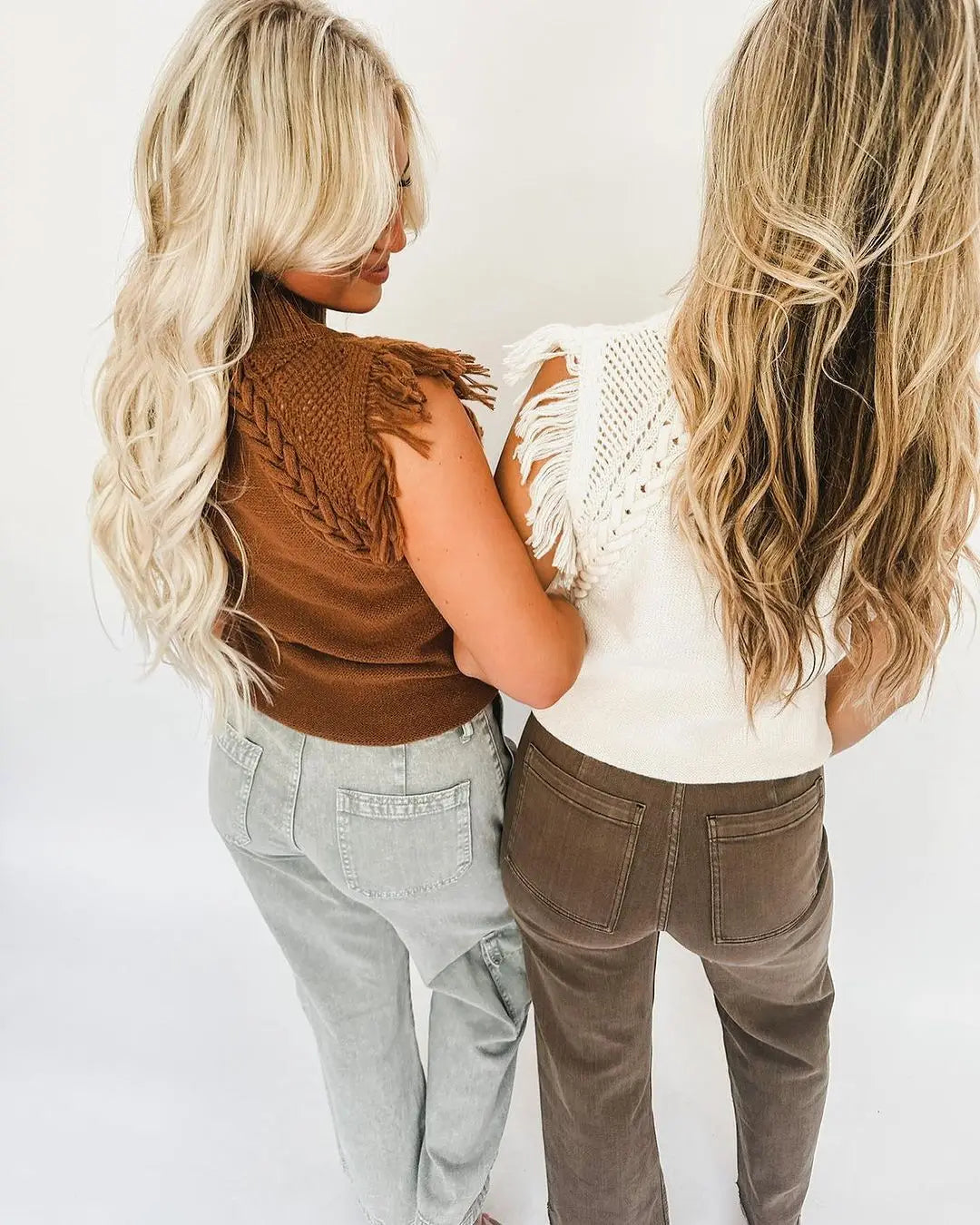 Two women stand back-to-back wearing a hand-woven fringed sleeveless knit sweater vest in white paired with dark brown corduroy pants and a brown knitted top with braided detailing paired with light-wash jeans.