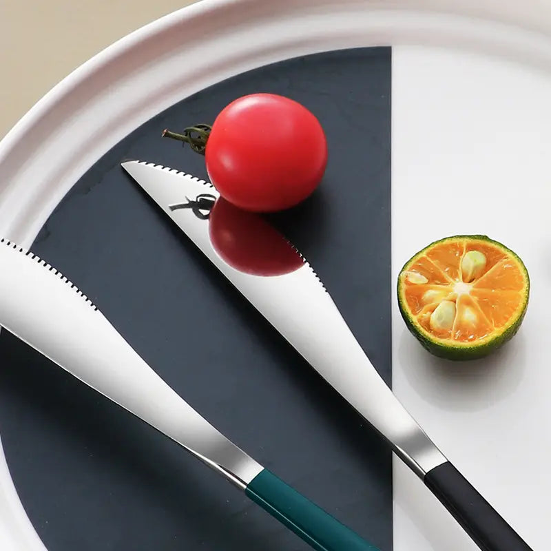 Two sleek stainless steel portable steak knives with serrated edges and contrasting teal and black handles rest elegantly on a modern plate beside fresh produce.