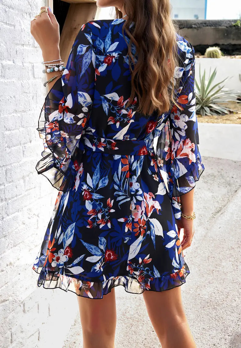 A navy blue women’s floral v-neck ruffle dress with waist tie featuring red and white blossoms on sheer fabric with ruffled sleeves and hem.