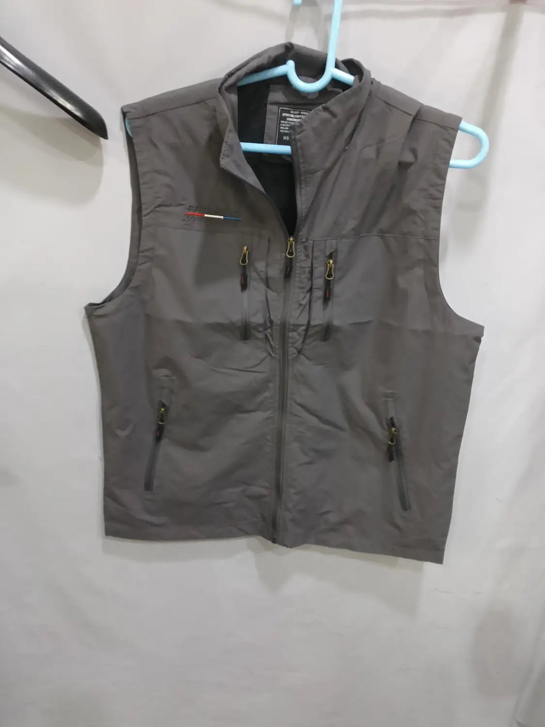 Chaleco utilitario con capucha y múltiples bolsillos para hombre confeccionado en nailon gris, con tiradores de cremallera negros y un pequeño logotipo rojo, blanco y azul en el lado izquierdo del pecho.