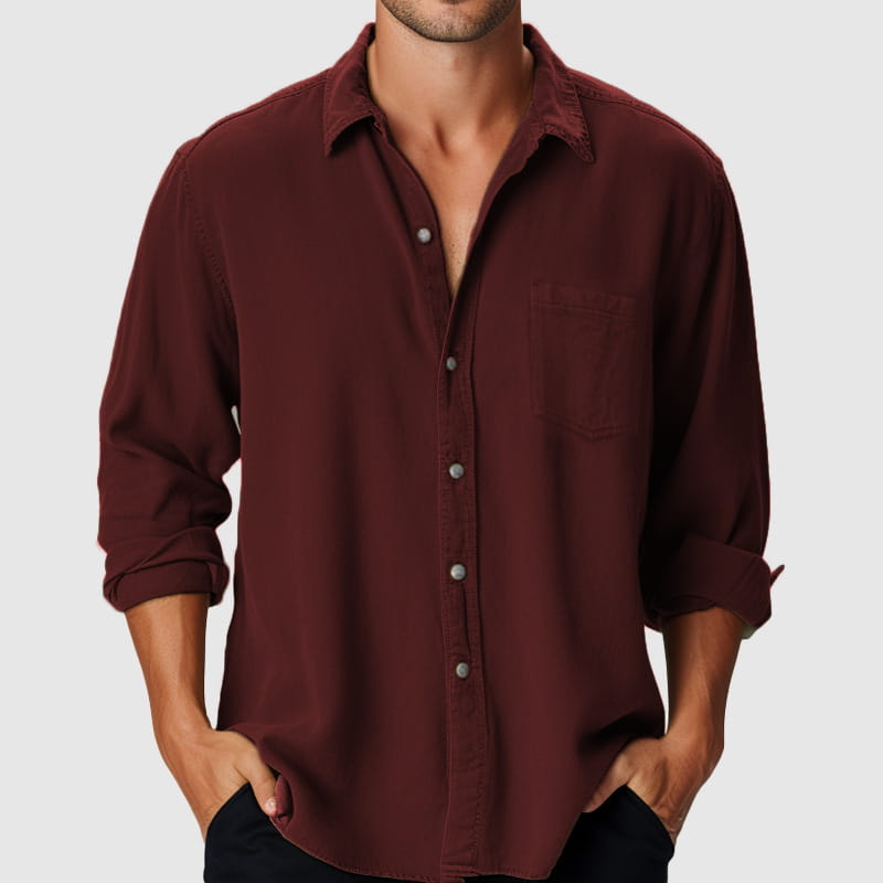 Ein bordeauxrotes Herren-Longsleeve-Freizeithemd am Strand aus leichtem Stoff mit Knopfverschluss vorne, umgekrempelten Bündchen und einer Brusttasche.