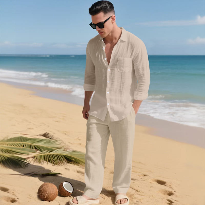 A man wearing a light beige men linen long sleeve shirt pants set made of breathable linen fabric standing on a sandy beach with rolled-up sleeves.