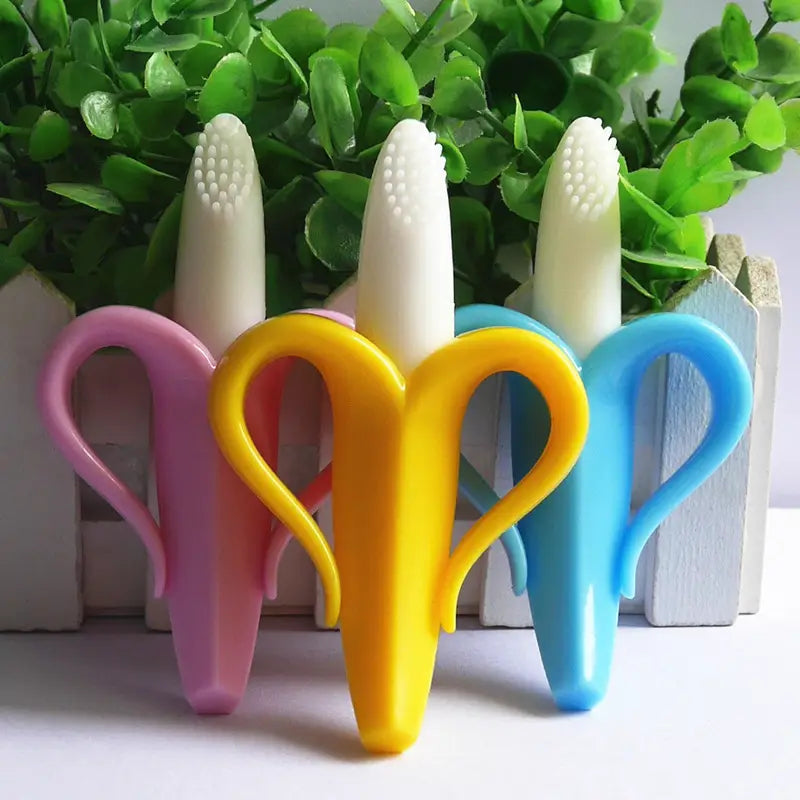Three colorful banana-shaped silicone baby teethers in pink, yellow, and blue with white bristle tips designed as a baby toothbrush toy.