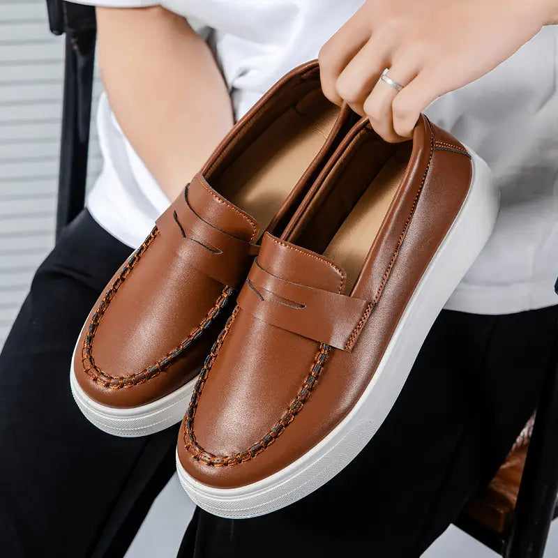 Mocasines marrones de cuero, tipo slip-on, con suela de goma blanca y costuras decorativas, diseñados para un uso casual.
