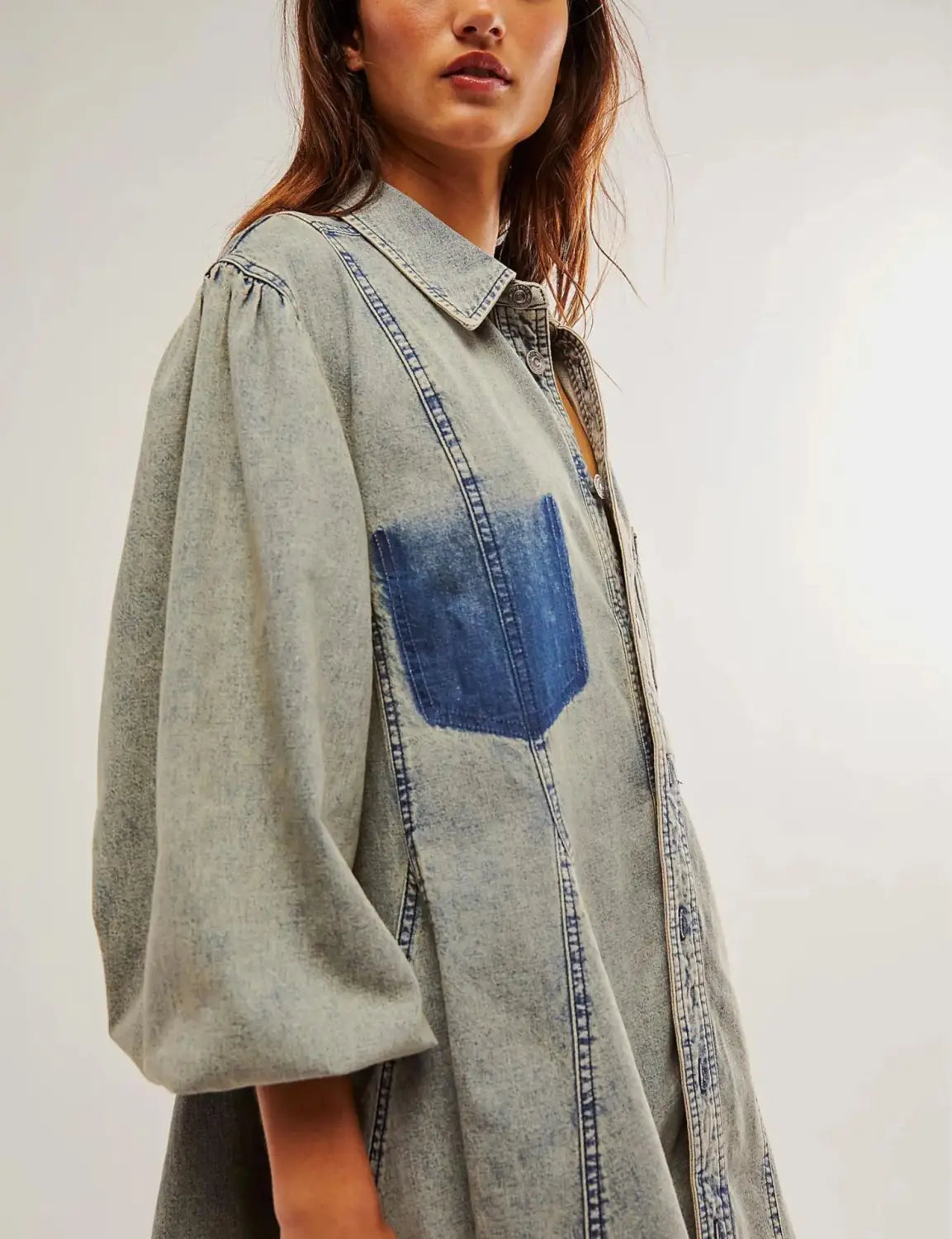 A light-wash denim shirt dress with lantern sleeves featuring a contrasting blue patch pocket and visible stitching details.