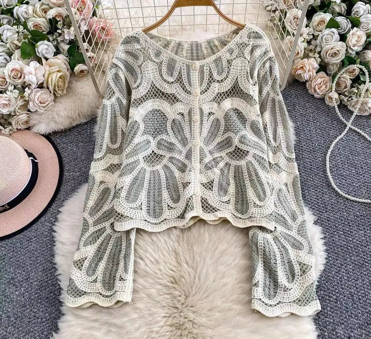 Ein zartes, elfenbeinfarbenes Häkel-Cardigan-Oberteil mit Blumenmotiv, locker sitzender Pulloverform, mit aufwendigen Spitzenblumenmustern und geschwungenen Abschlüssen, präsentiert auf einem Holzkleiderbügel.