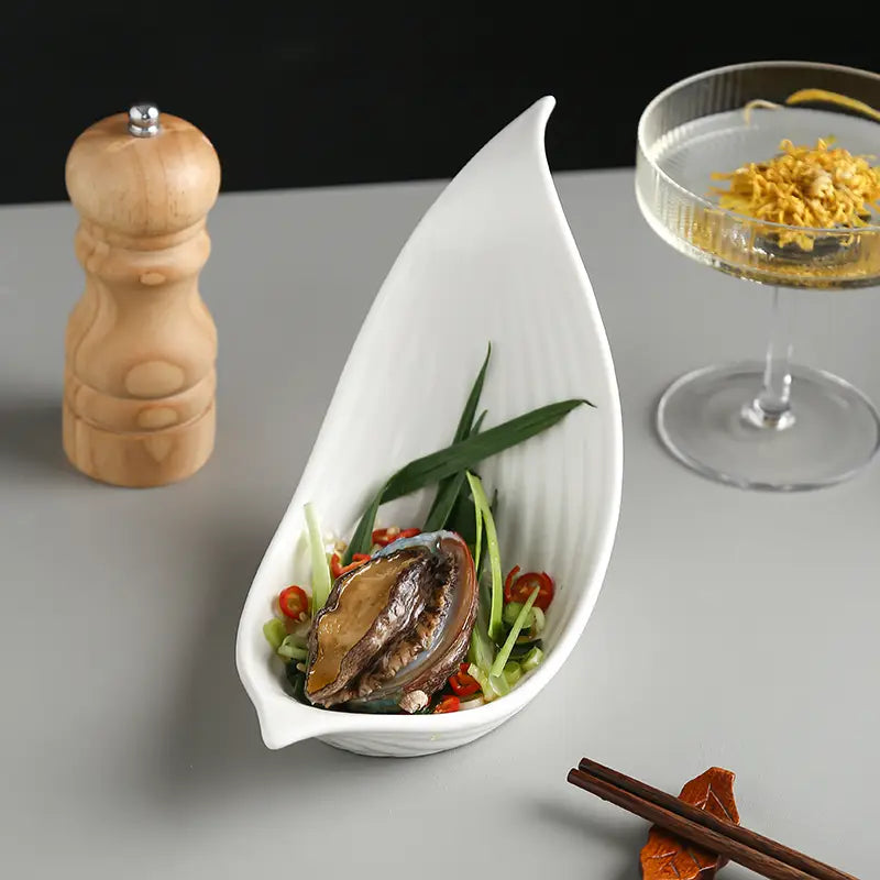 White ceramic leaf-shaped serving dish with a glossy finish holding an abalone and garnishes in a white leaf ceramic plate irregular serving dish.