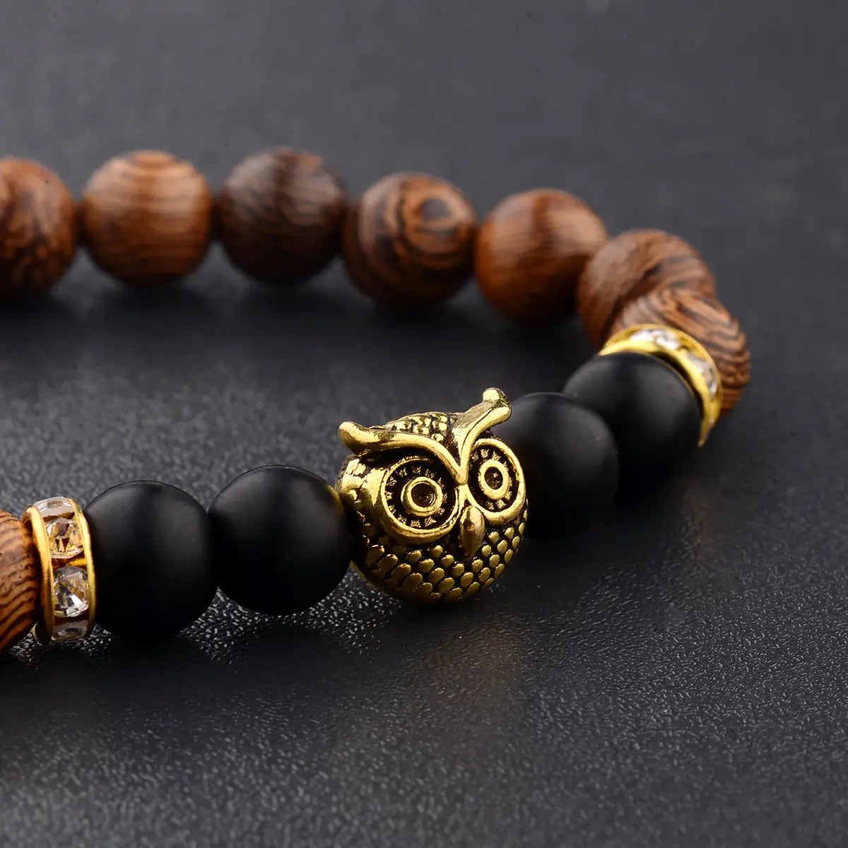 A gold-toned owl charm adorns a crystal owl charm wood and onyx beaded bracelet featuring dark brown wooden beads black matte beads and crystal-encrusted metal spacers.