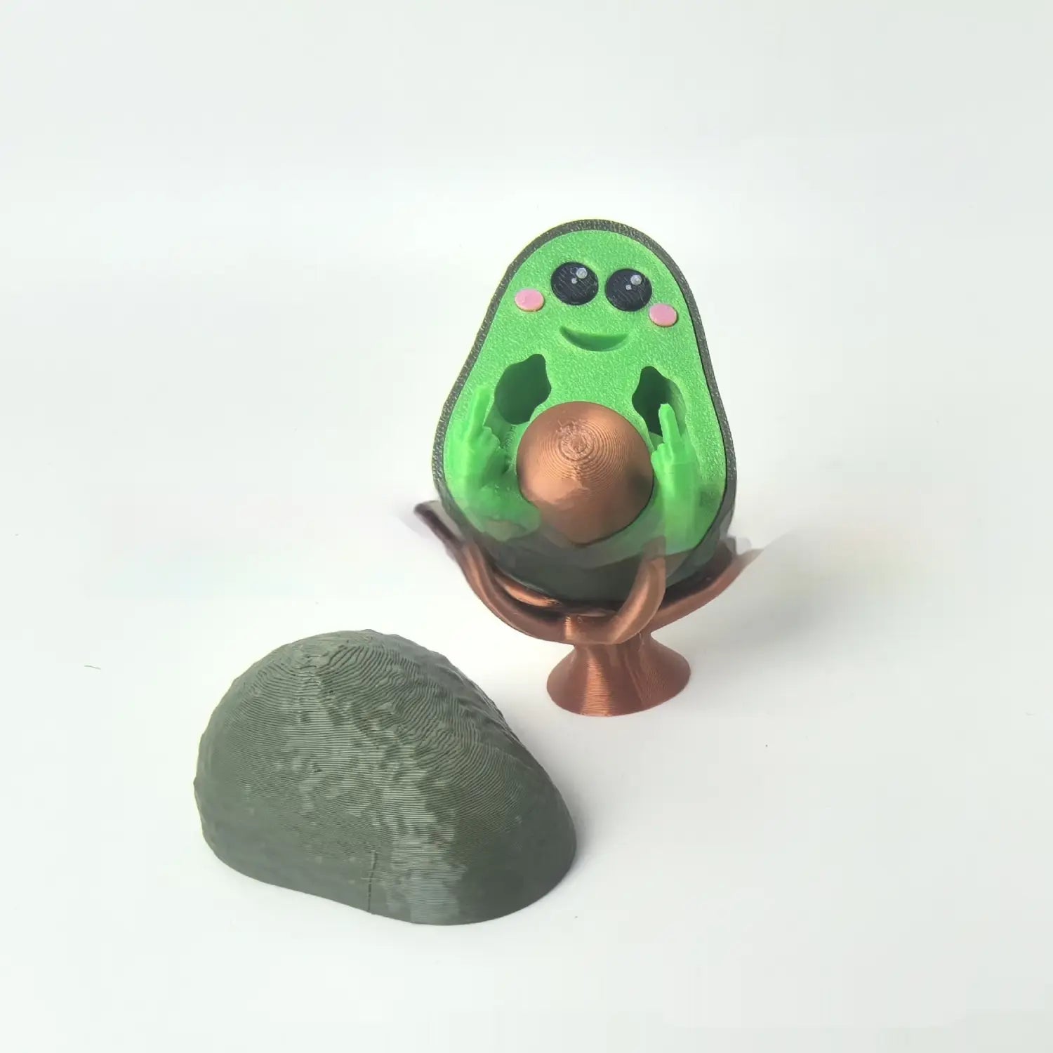 Green avocado-shaped 3d printed desk toy phone stand with black eyes, pink cheeks, smiling face, and copper-colored sphere on pedestal base measuring 6.5cm.