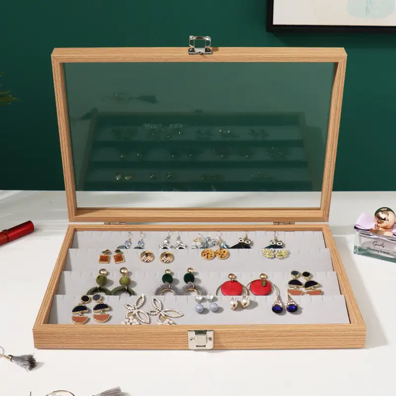 Rosewood jewelry display box with glass lid and silver-toned latches in light wood finish featuring internal compartments holding various earrings.