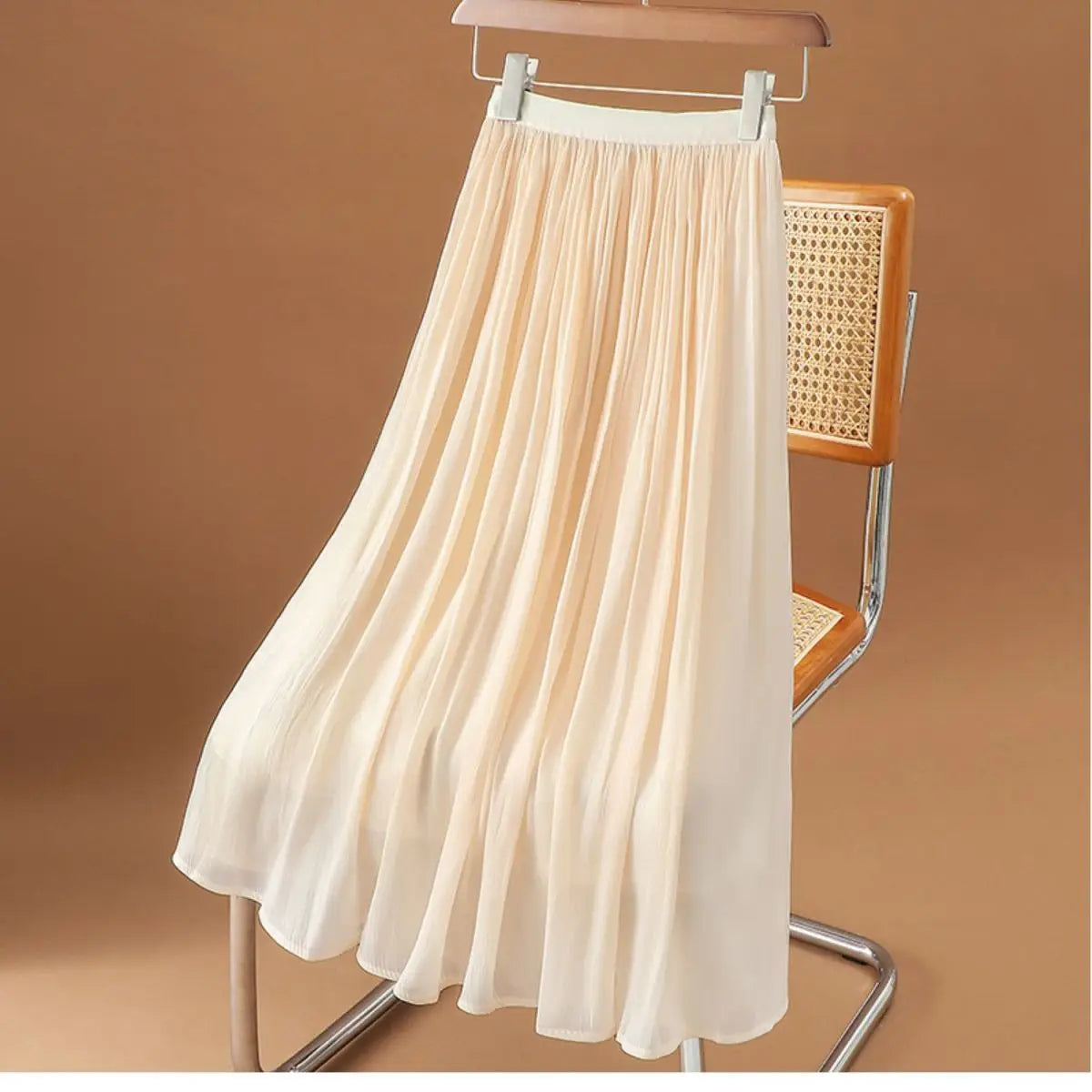 Ein fließender cremefarbener irrisierender Chiffon-Faltenrock aus leichtem Gewebe, der elegant an einem Holzkleiderbügel hängt.