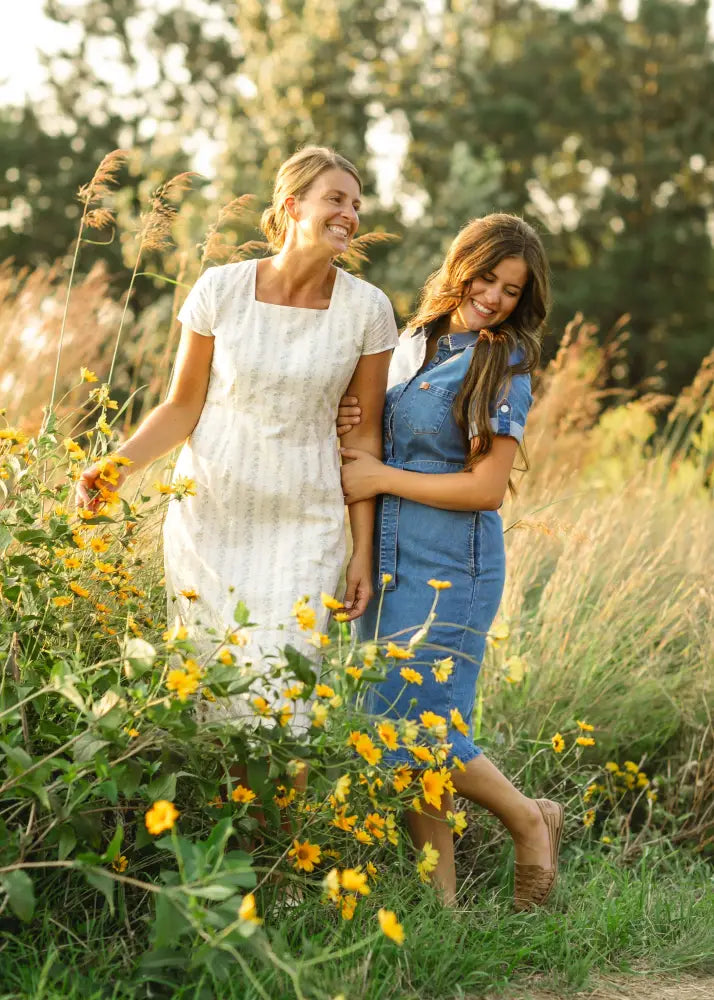 Two women stand in a field of yellow flowers wearing a white dress and an ada denim button front midi dress with belt