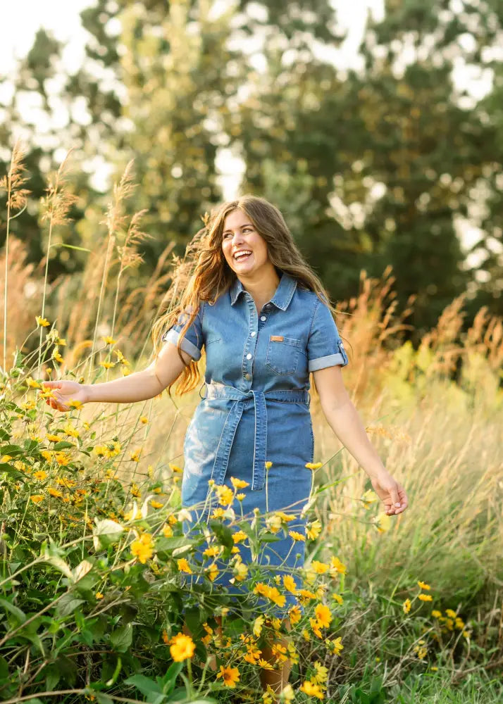 A woman standing in a field wearing an Ada denim button front midi dress with belt in blue denim with short sleeves