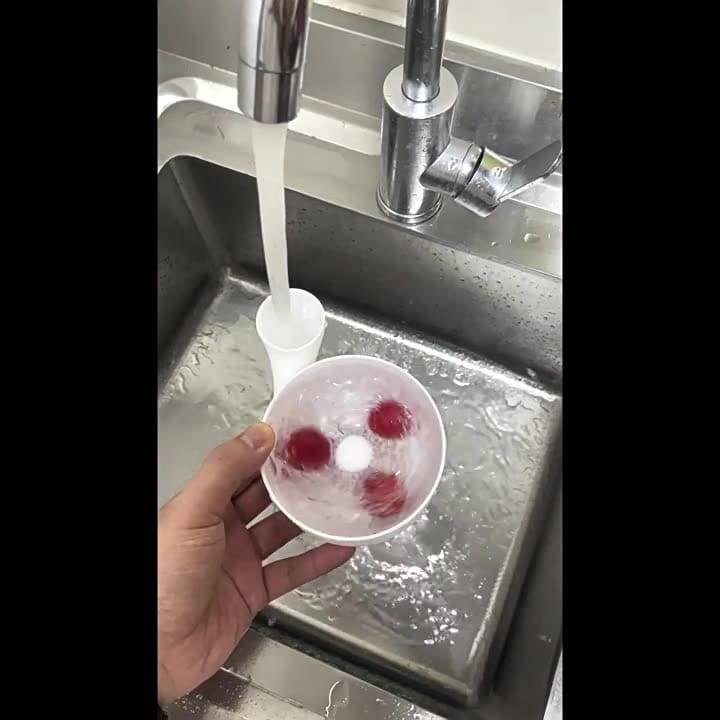 Bol de lavage de fruits en plastique blanc avec fruits sphériques rouges flottant à l’intérieur, maintenus sous l’eau courante dans un évier en acier inoxydable, dans le cadre d’un ensemble de presse-agrumes.