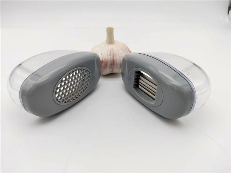 Presse-ail en plastique gris et transparent avec lames en acier inoxydable, comprenant un côté râpe perforé et un côté lame écraseuse pour les gousses d’ail.