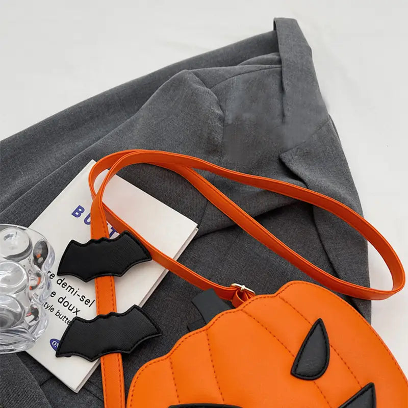 Orange pumpkin-shaped crossbody bag with black bat embellishments, stitched details, and detachable charm designed for festive use.