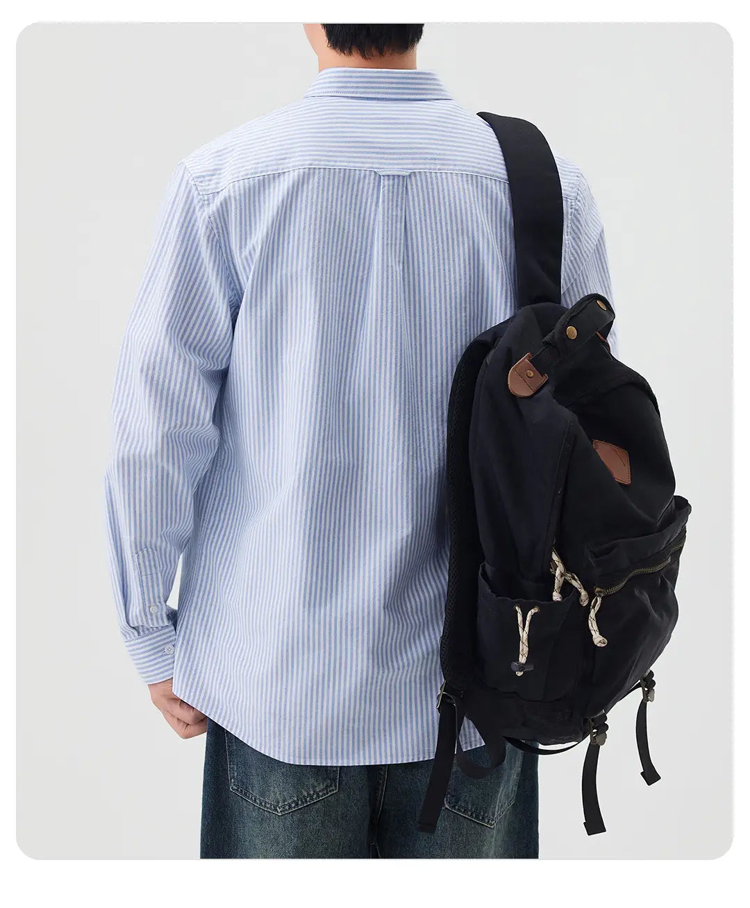 Mochila negra de lona con detalles en cuero marrón y tiradores con cierre con cremallera, mostrada por el lado posterior, en combinación con la camisa de manga larga abotonada con rayas para hombres