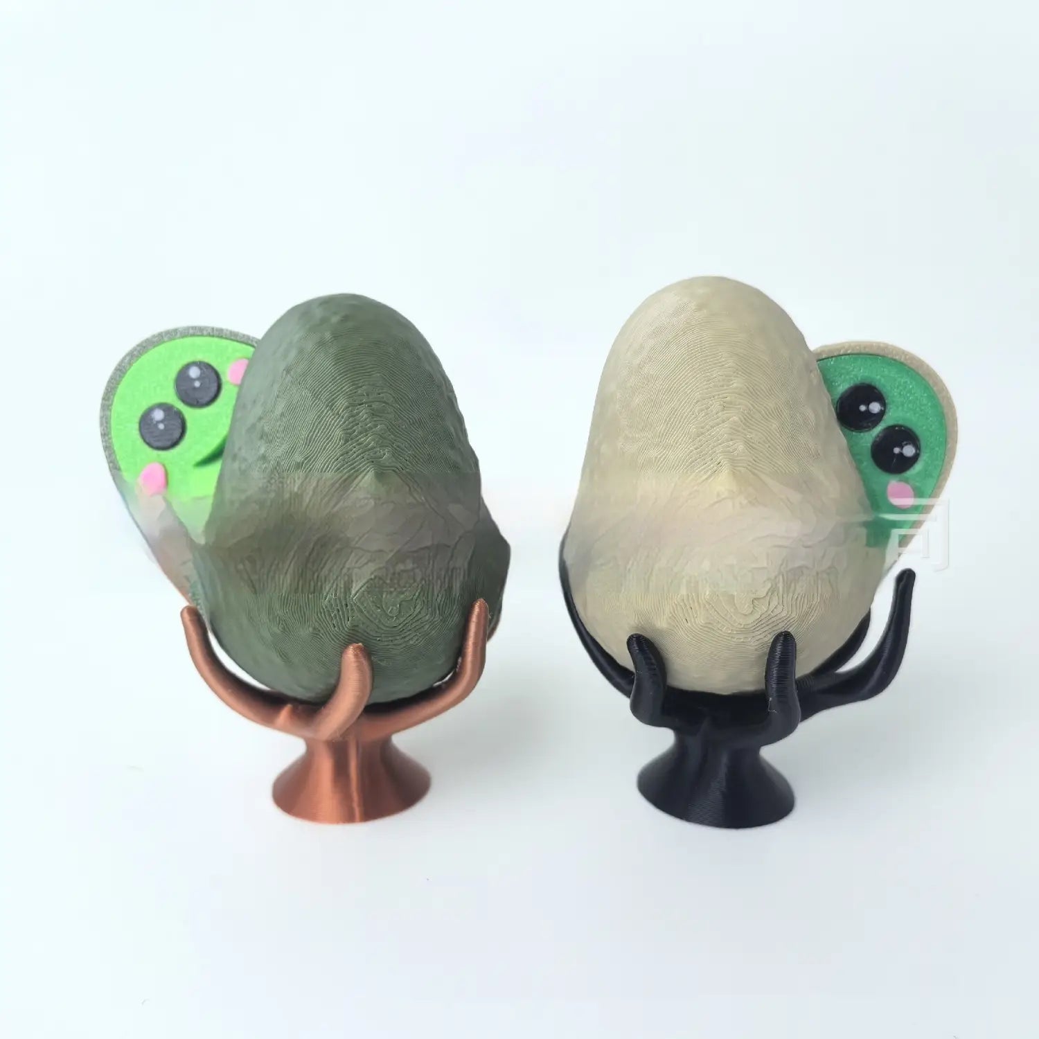 two 3d-printed avocado phone stands featuring green cartoon faces peeking from behind egg-shaped holders supported by stylized hand-like stands for desk decoration and utility