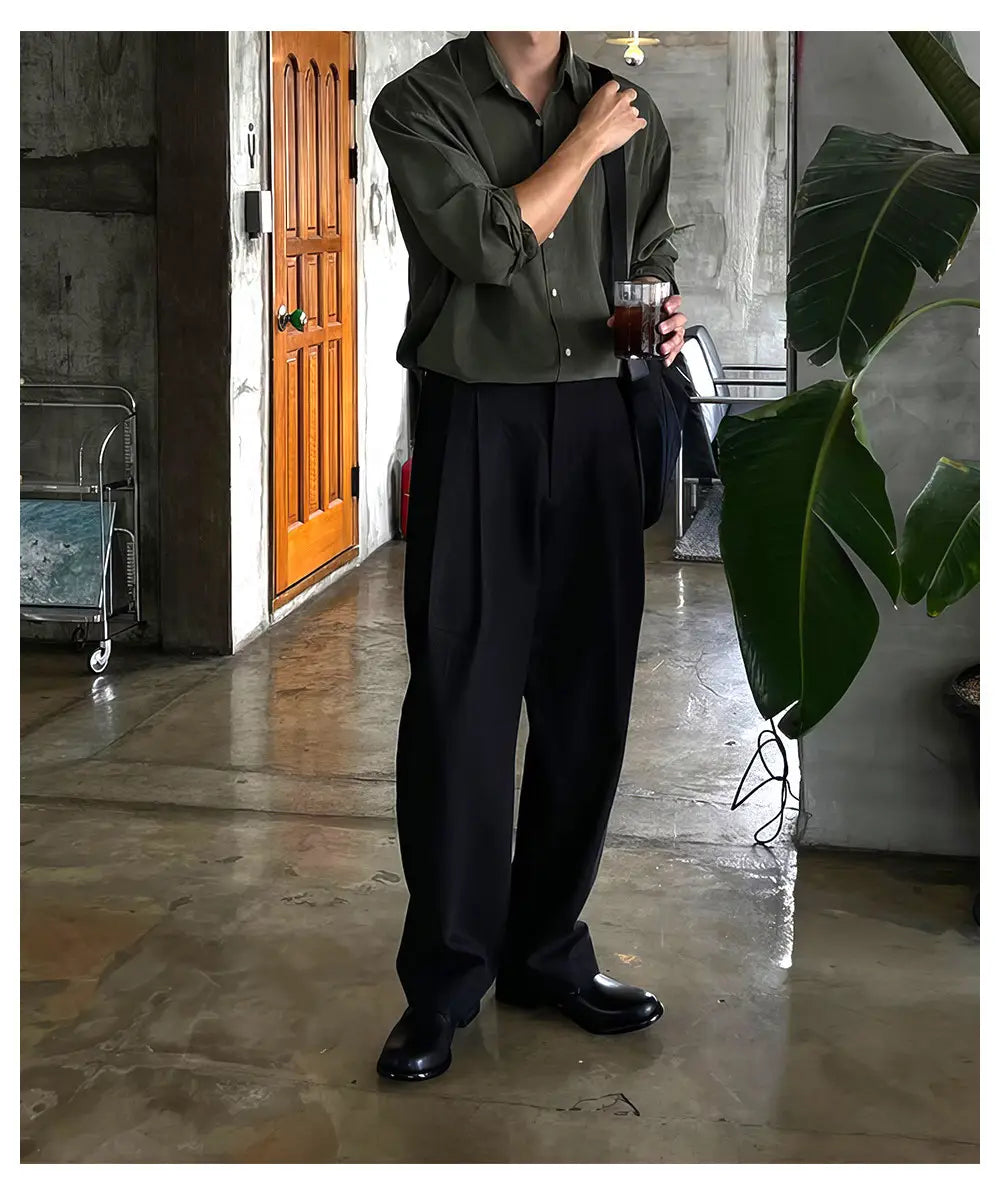 Pantalones de vestir anchos plisados color gris oscuro para hombre, combinados con una camisa abotonada verde oliva con mangas arremangadas y una textura sutil.