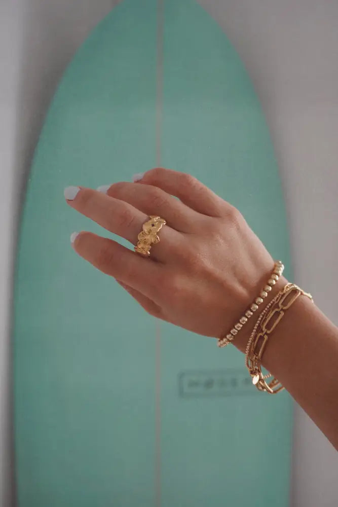 A delicate gold tropical flower statement ring on the finger with matching gold chain and beaded bracelets