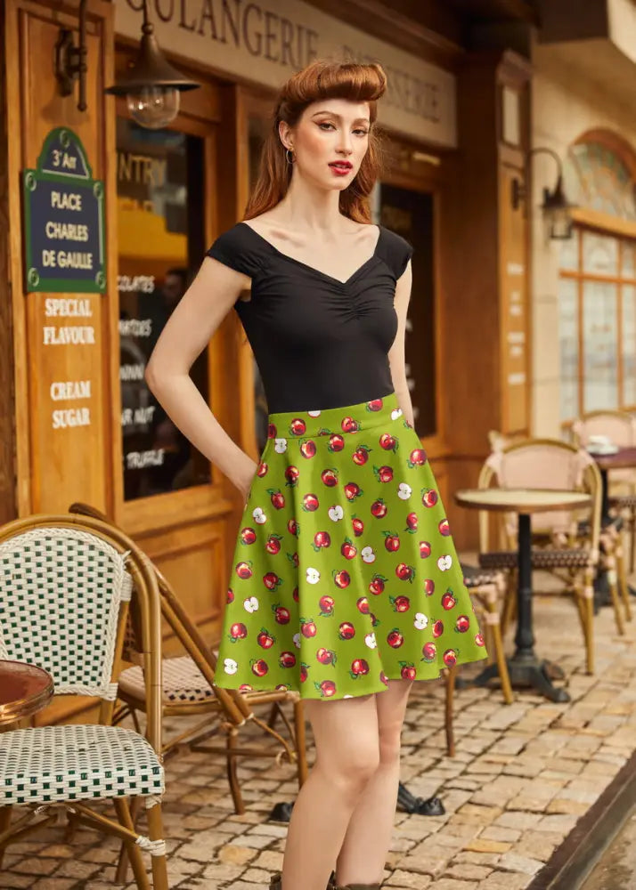 A vibrant green pleated skater skirt featuring a playful red and white apple print in a-line style