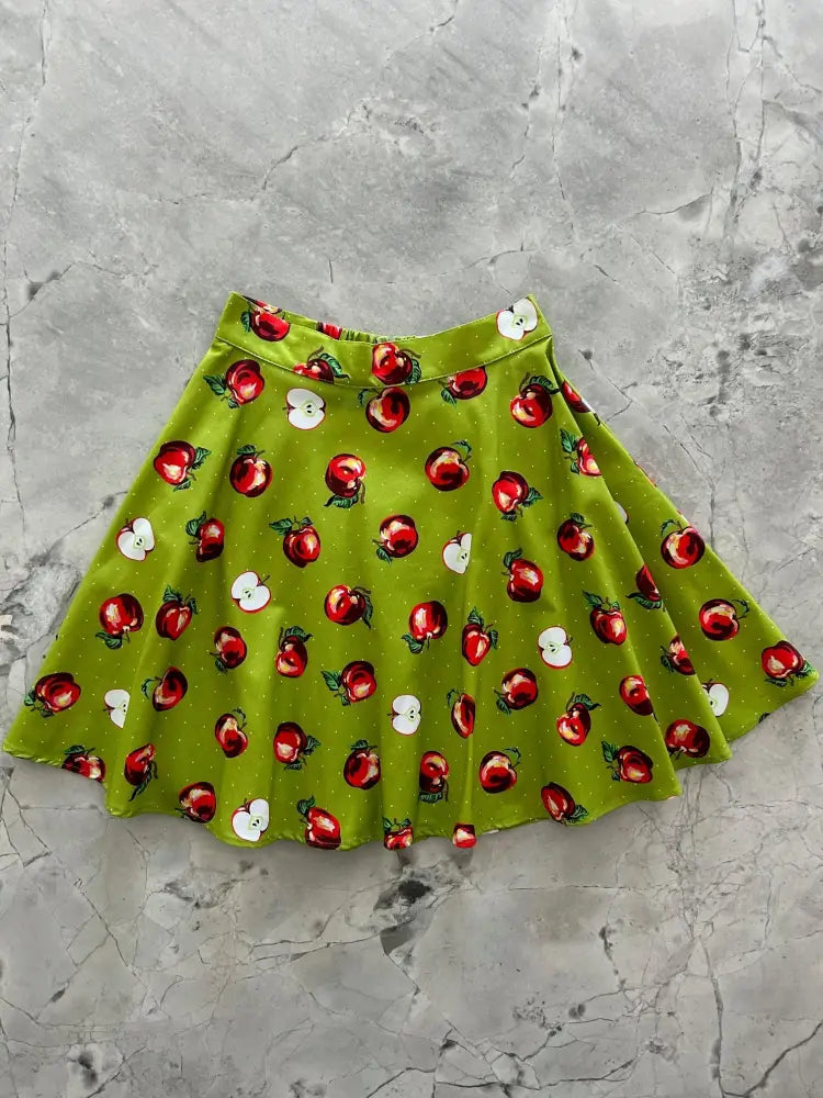 A vibrant lime green pleated skater skirt featuring a playful red apple and white apple slice print