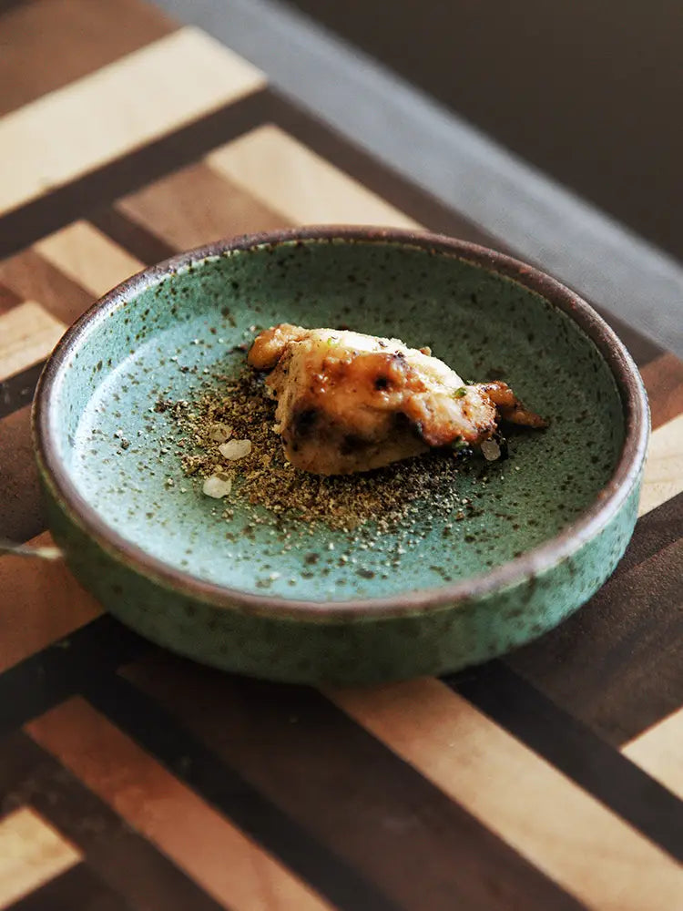 A rustic green ceramic sauce dish with speckled glaze and dark brown rim holding a single seared piece of food garnished with coarse seasoning designed as a Japanese dipping bowl.