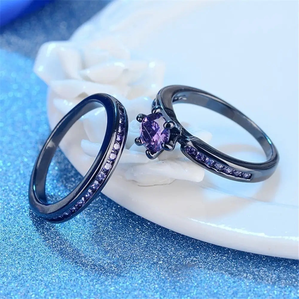 Black tungsten and purple gemstone wedding band set featuring a black ring with a central purple gemstone surrounded by smaller stones and one band adorned with sparkling accents.
