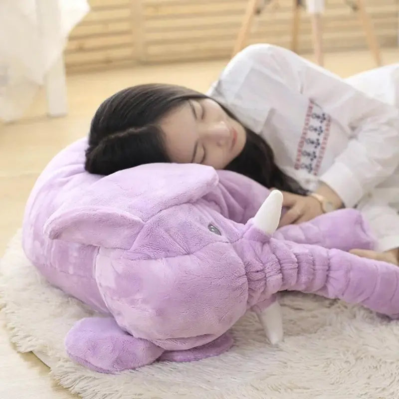 Coussin en peluche lavande représentant un éléphant, avec défenses blanches et tissu souple à texture délicate, conçu comme un coussin d’animal en peluche pour les enfants.