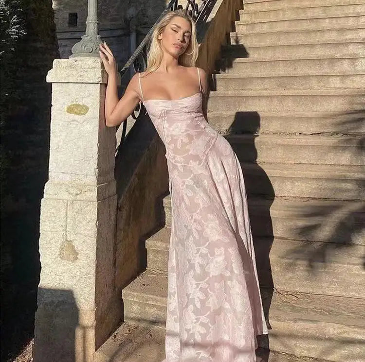 Une robe de soirée maxi sans dos en dentelle florale rose pâle, fluide, avec des motifs délicats et de fines bretelles spaghetti, illustrée depuis la face arrière.