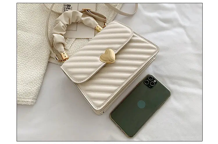 Cream-colored quilted heart clasp crossbody bag with ruched handle featuring a gold heart-shaped clasp and matching adjustable strap.