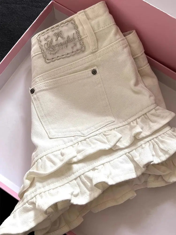 Falda de mezclilla color marfil con dobladillo con volantes y parche bordado, presentada como pantalones cortos de mezclilla con volantes color crema de cintura alta.