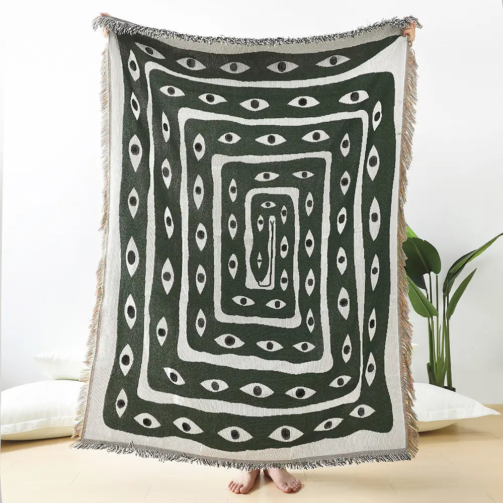A dark green and white woven cotton throw blanket featuring a repeating pattern of stylized eyes within concentric rectangular frames and fringed borders.