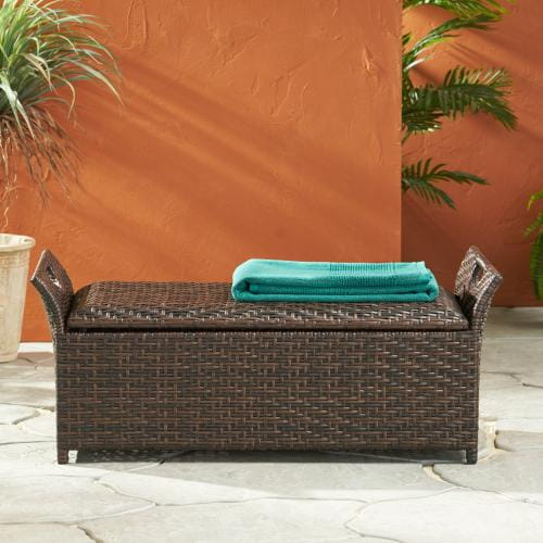 Brown woven wicker storage bench with matching lid and handle ends topped by a folded teal towel designed for outdoor seating.