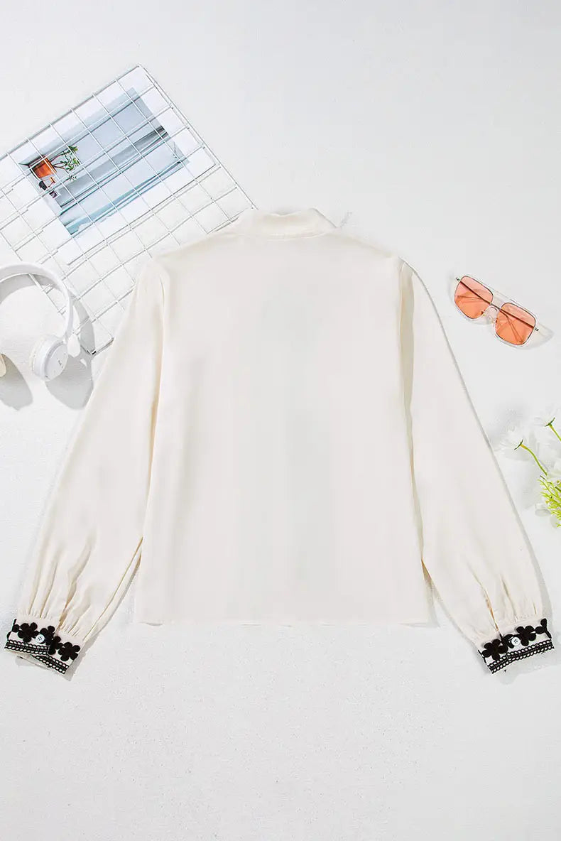 Cream-colored long sleeve blouse with black floral embroidery at the cuffs and delicate lace trim suitable for elegant wear.